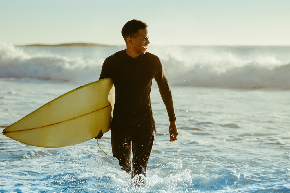 Smiling young man with surfboard on the beach. African male coming out of the ocean after water surfing.